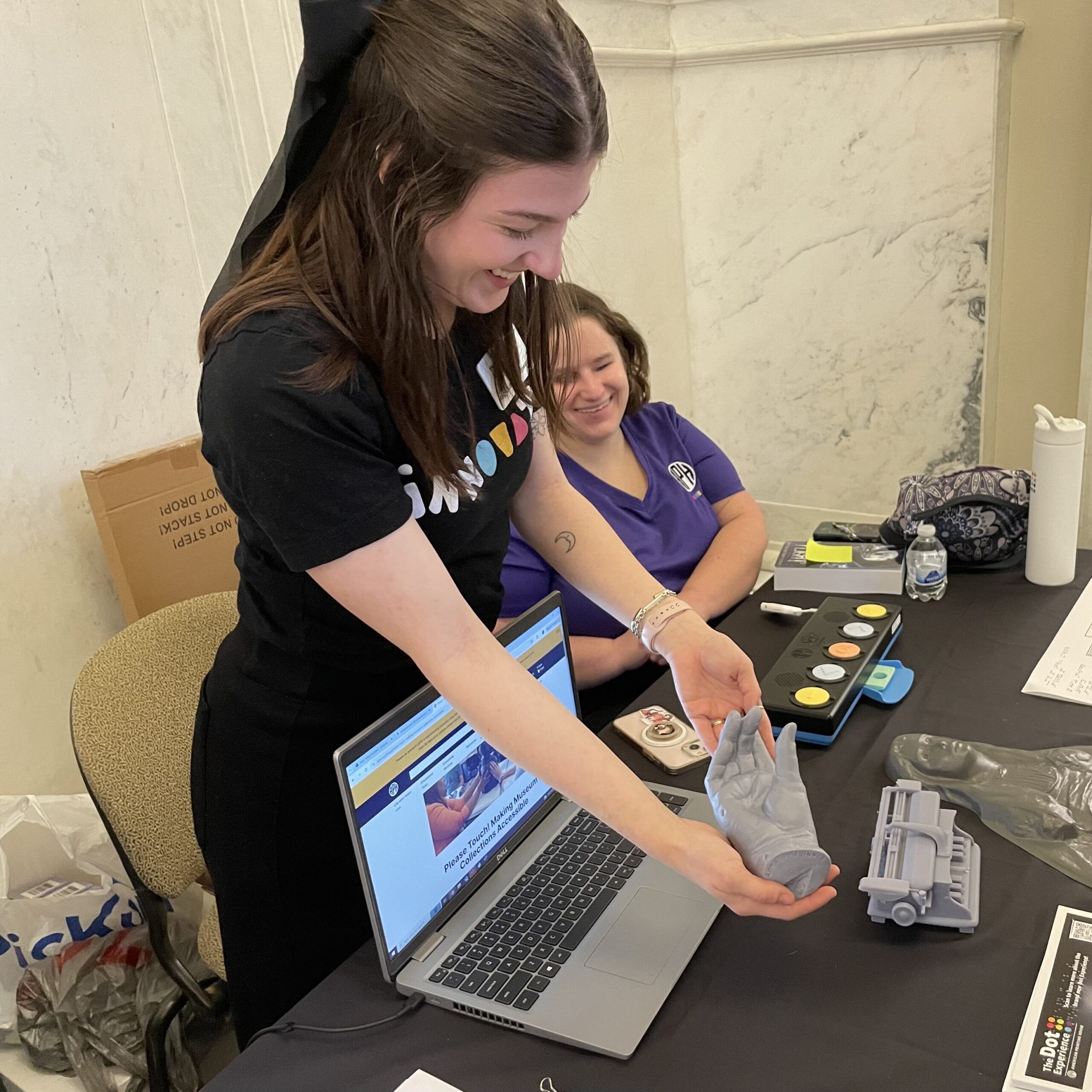 Two people behind a table, one sitting and one standing, smile as they show a grey cast of a hand to a person standing on the other side of the table. A laptop, a Joy Player, a tactile map, and several other prototypes are visible on the table.