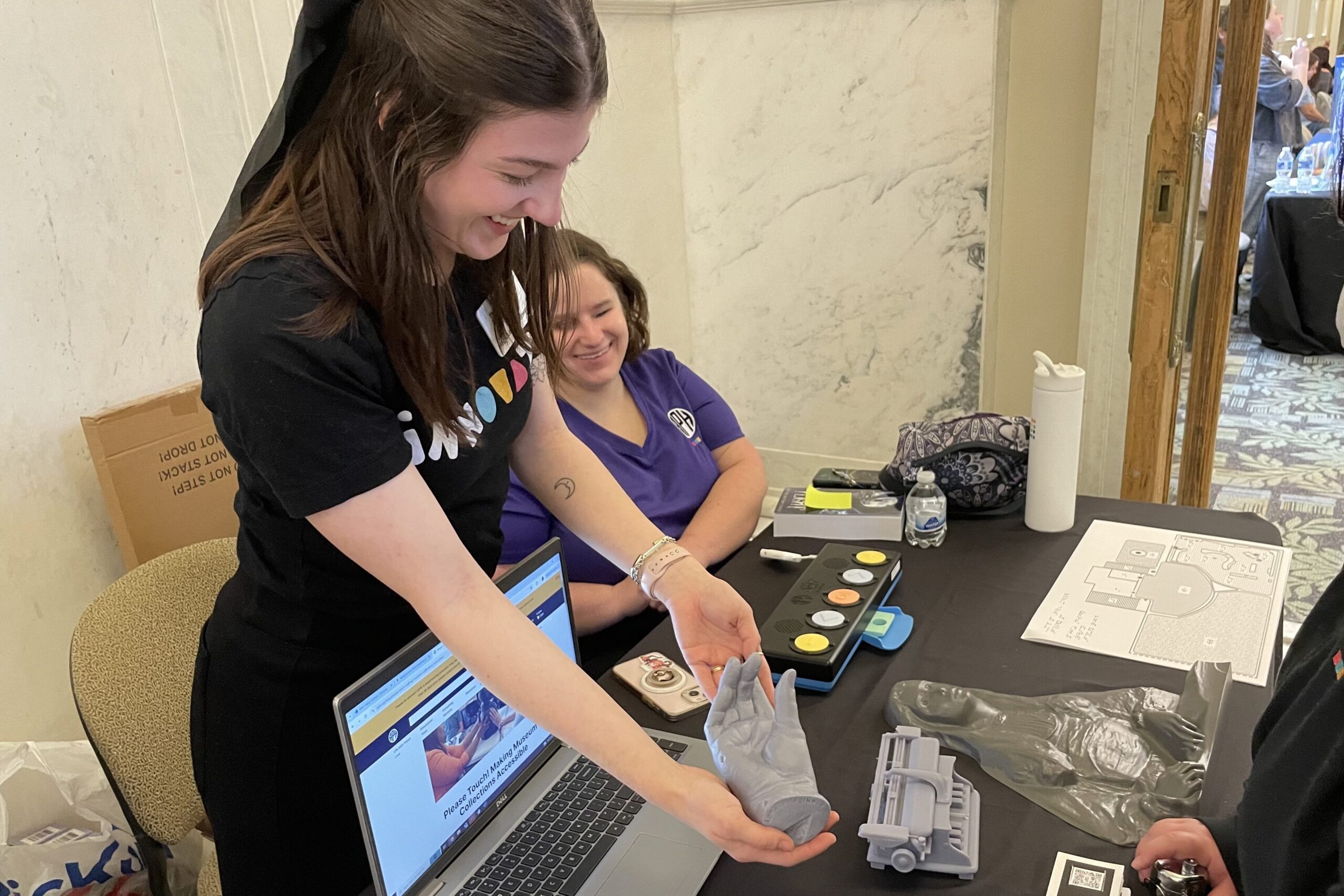 Two people behind a table, one sitting and one standing, smile as they show a grey cast of a hand to a person standing on the other side of the table. A laptop, a Joy Player, a tactile map, and several other prototypes are visible on the table.