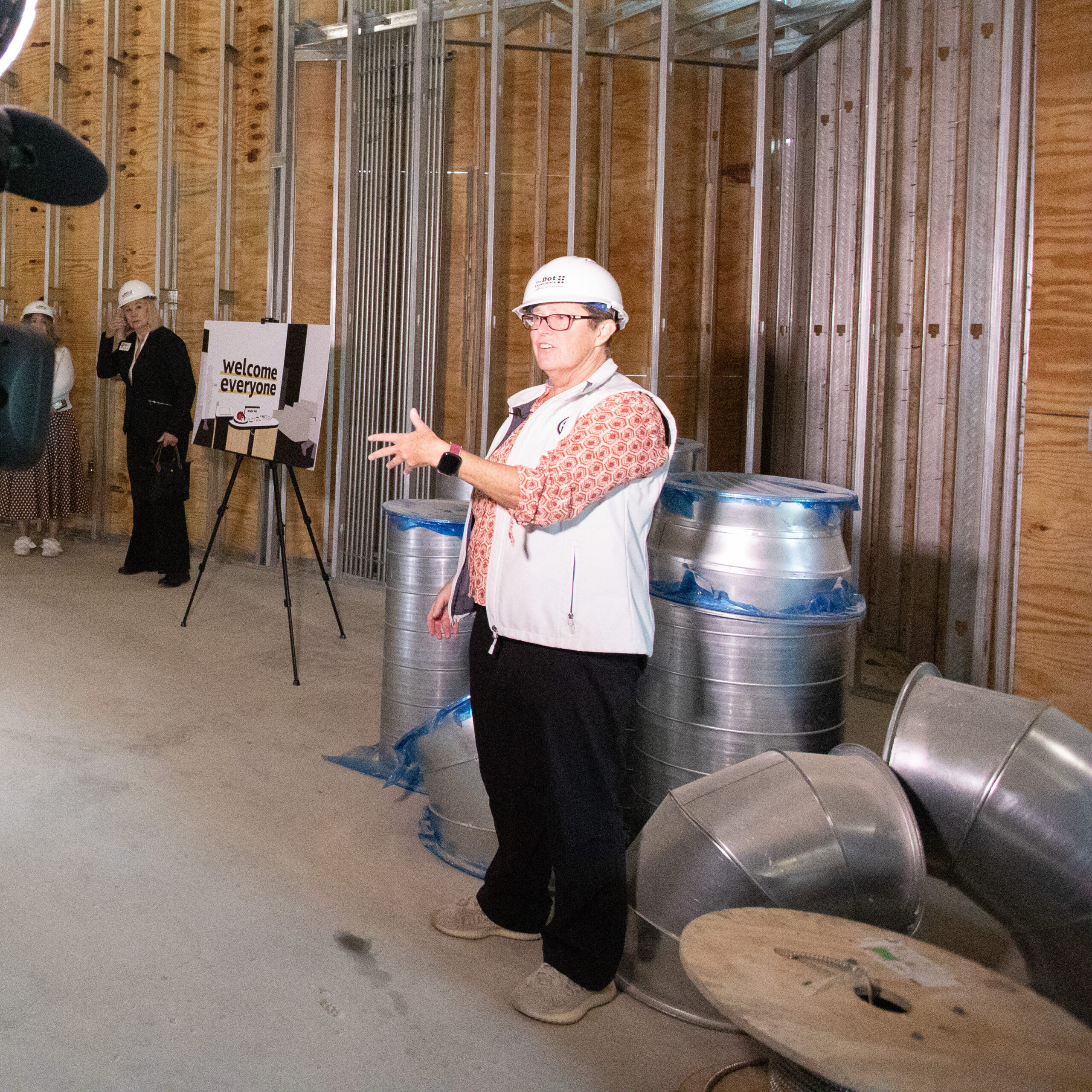Cameraman in hard hat filming a woman in hard hat speaking in front of a partially constructed room with metal framed walls and a tall ceiling.