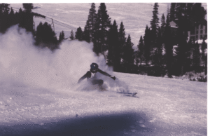 A skier makes a turn on a snowy mountainside with pine trees, whipping up snow behind him
