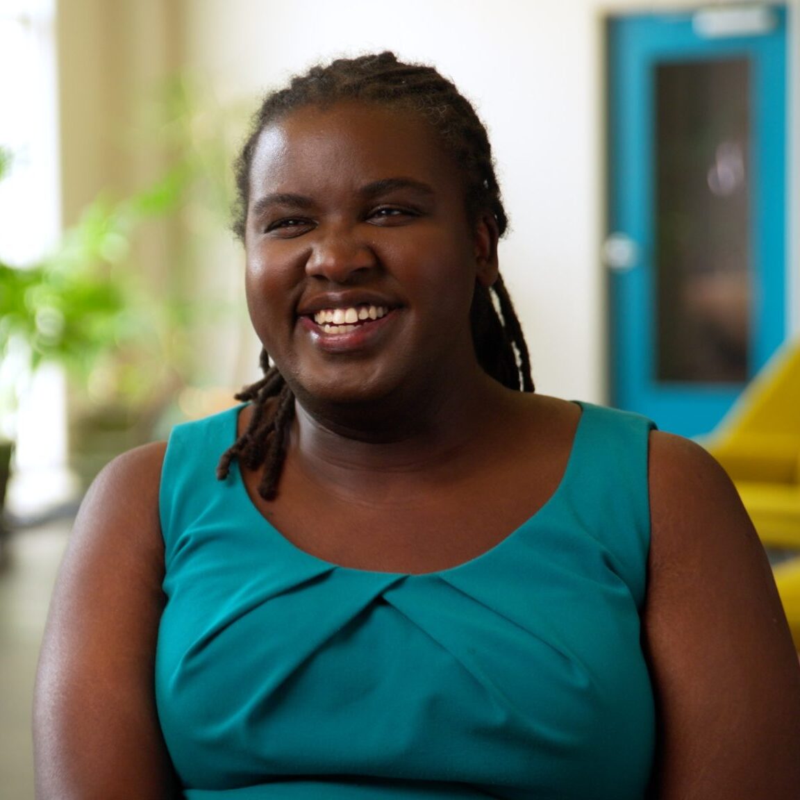 Denna Lambert smiled in a well-lit room where plants and a yellow sofa are visible behind her.