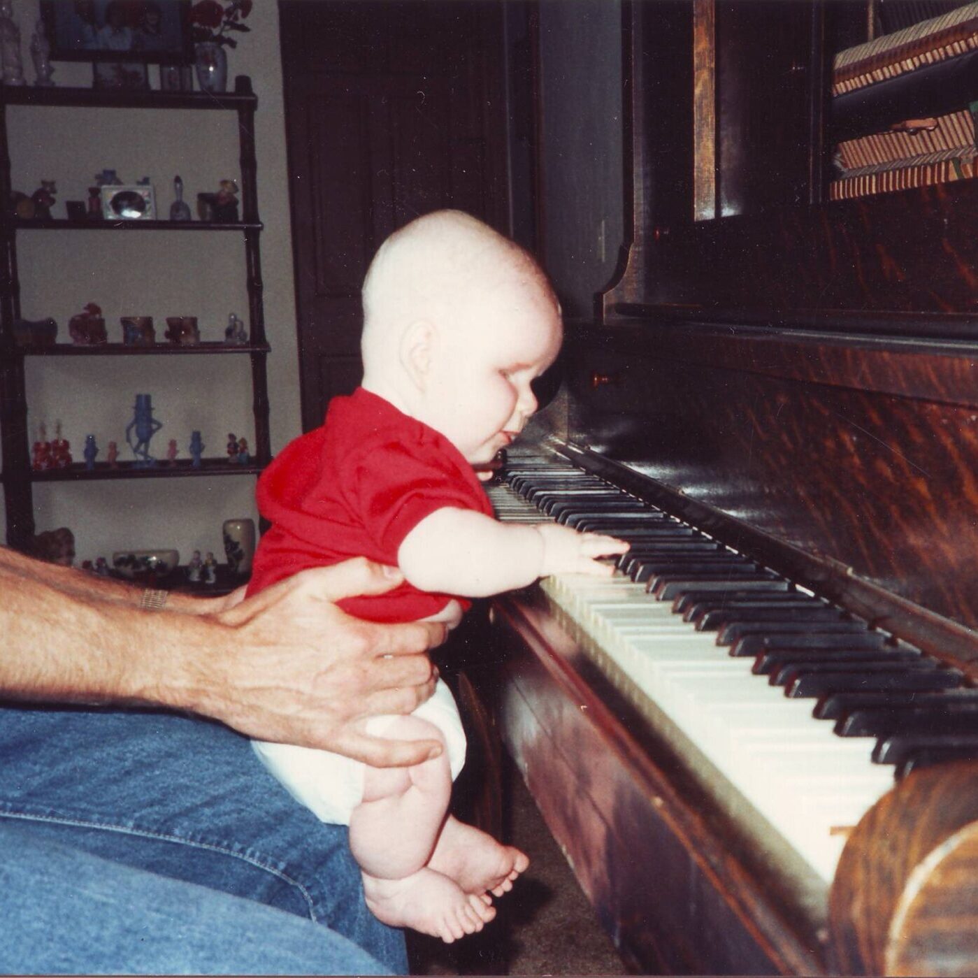 A young Patrick, around the age of 9 months, is being held up in front of the piano where he plays the keys.
