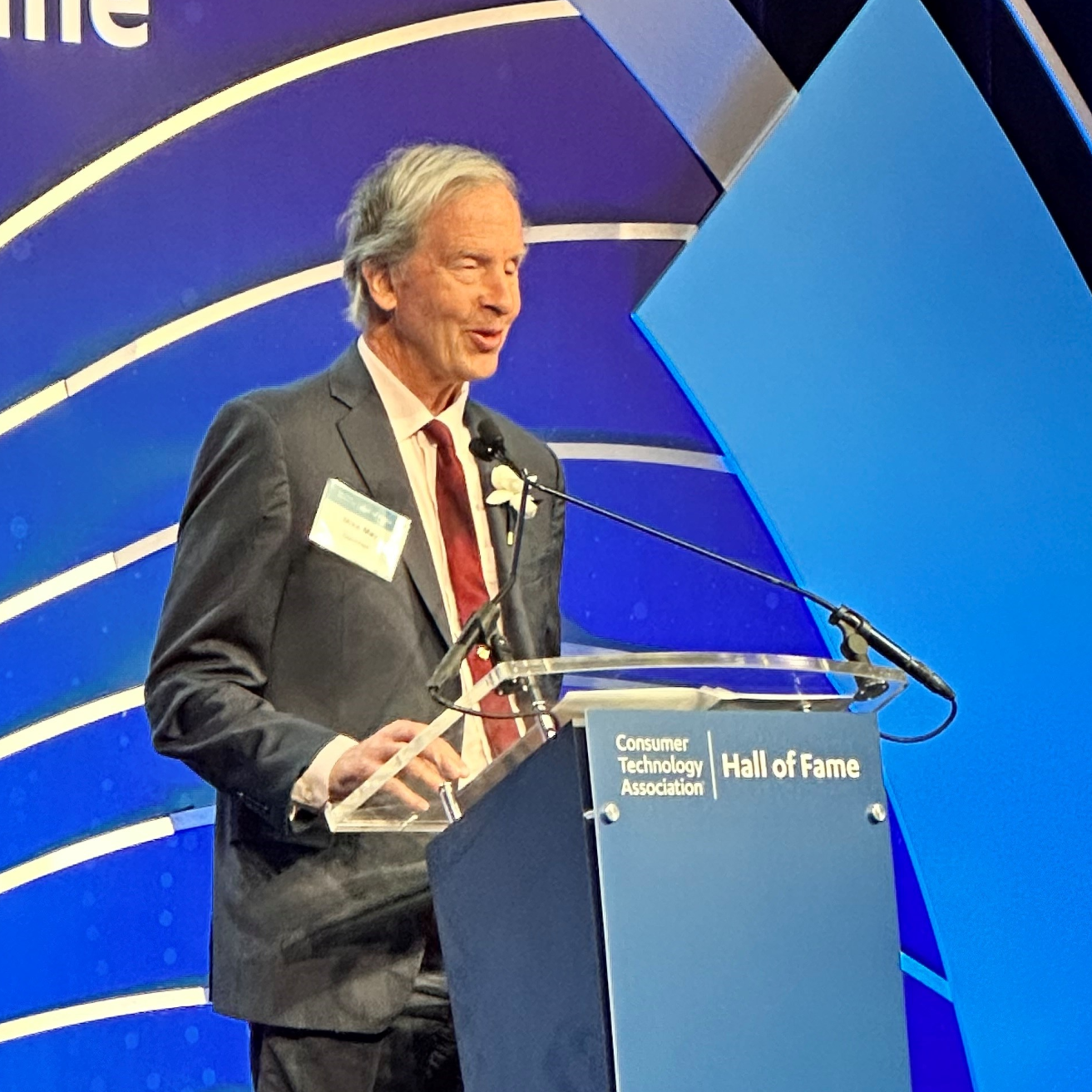 Mike May wears a suit as he speaks from a podium with the words 