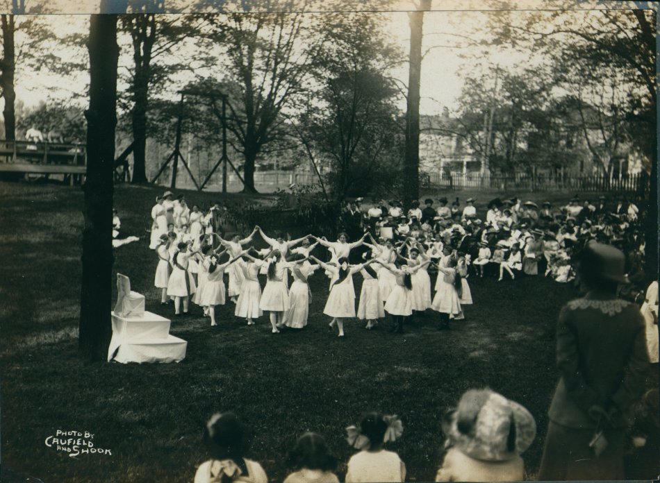 Girls folk dancing in a circle in front of a seated crowd, May 1, 1913.
