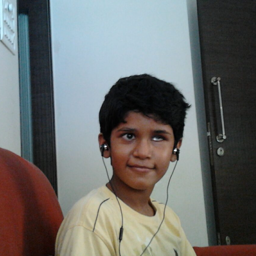 A young Bhavya, wearing a yellow t-shirt and sitting on an orange couch, smiles at the camera. He is wearing wired black headphones.