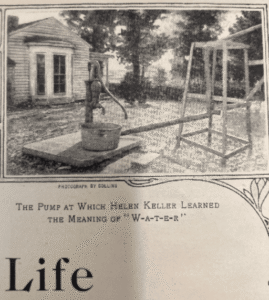 Newspaper image of water pump in front of house. The image has been cropped from the original and there is type set underneath that says "THE PUMP AT WHICH HELEN KELLER LEARNED THE MEANING OF "W-A-T-E-R".