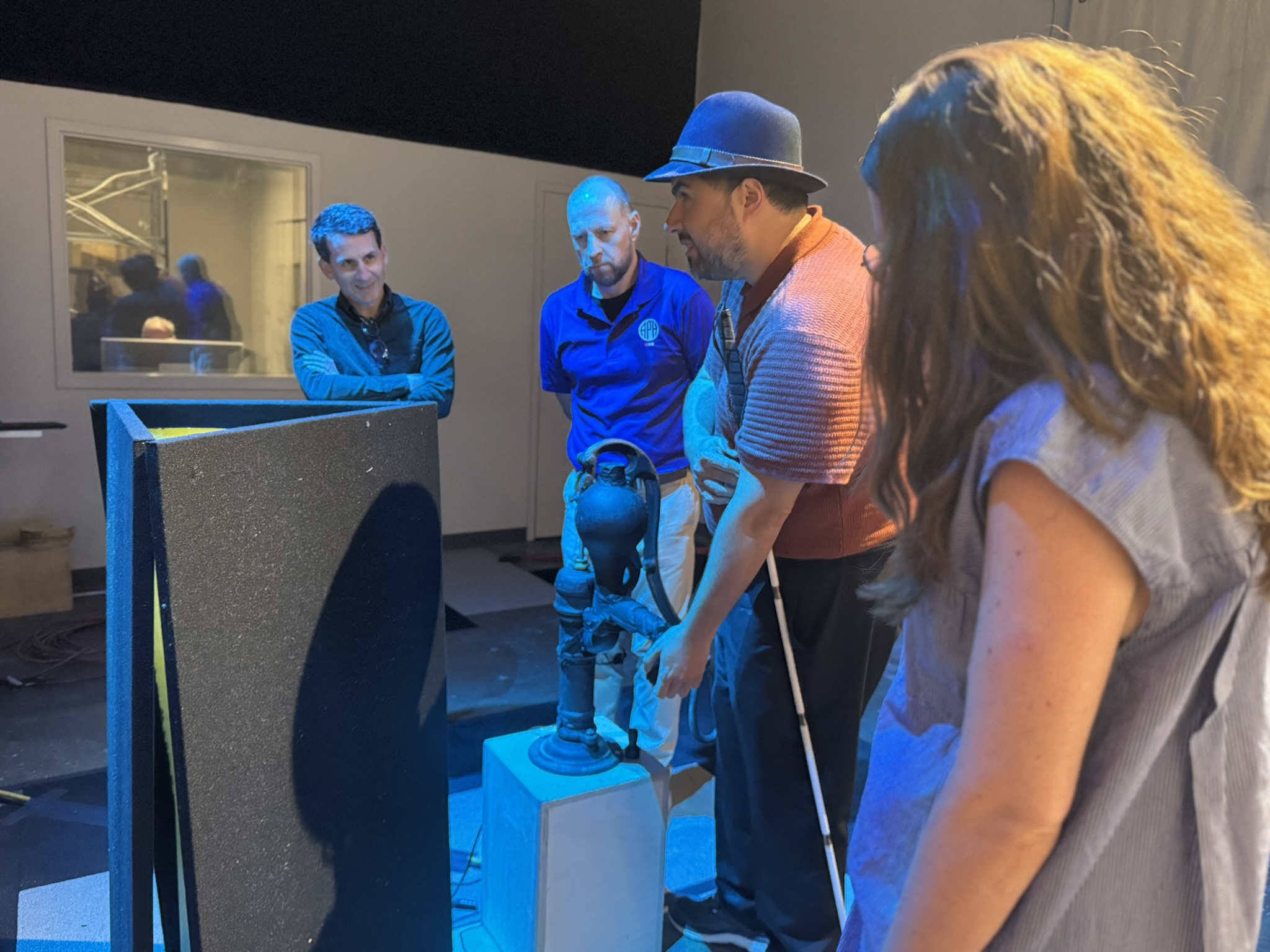 Four adults, one with a cane, stand around the 3-D water pump and feel the exhibit.