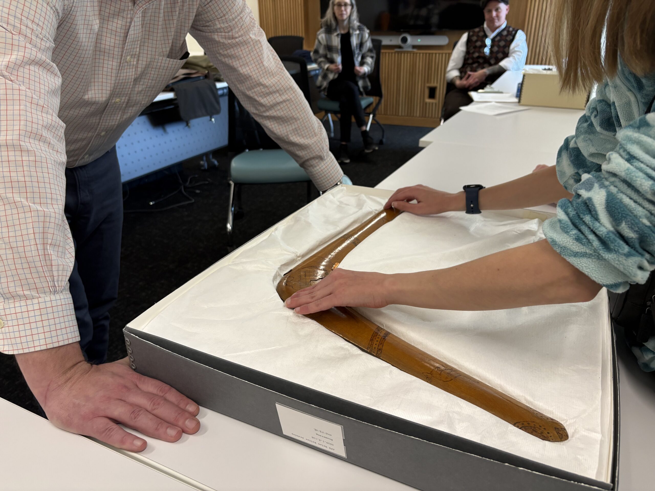 "A member of the prototyping team reaches out to touch a large wooden boomerang laying in an open box on a table. "