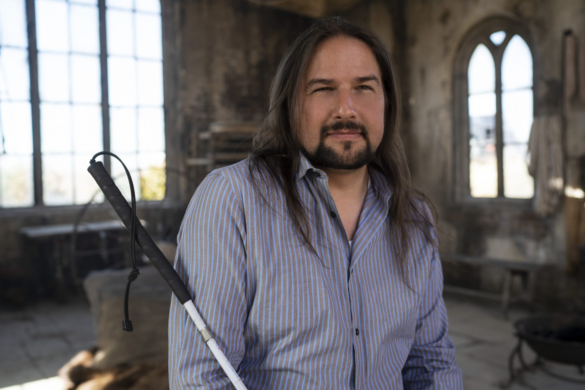 A man with long brown hair and a beard looks into the camera. He is wearing a blue shirt, holding a white cane, and is seated in front of large windows
