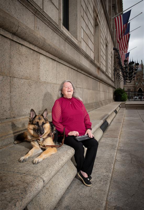 Woman wearing red shirt and black pants is leaning against a textured concrete wall. A German Shephard guide is sitting next to her and both are looking the camera.