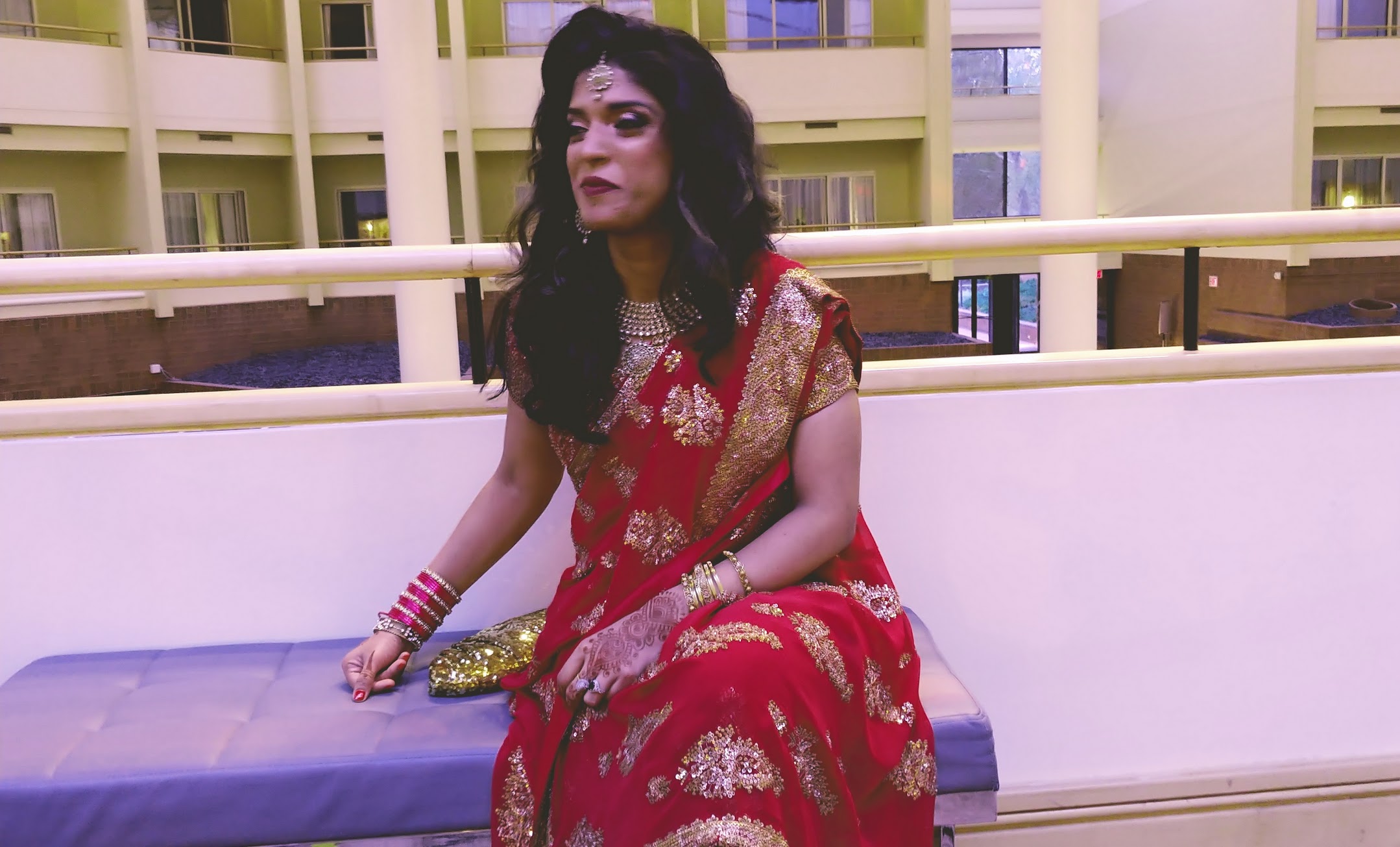A woman with long dark hair is seated looking away from the camera. The woman is wearing a traditional Indian dress called a Sari. It is flowing around her body and is red with gold decorations