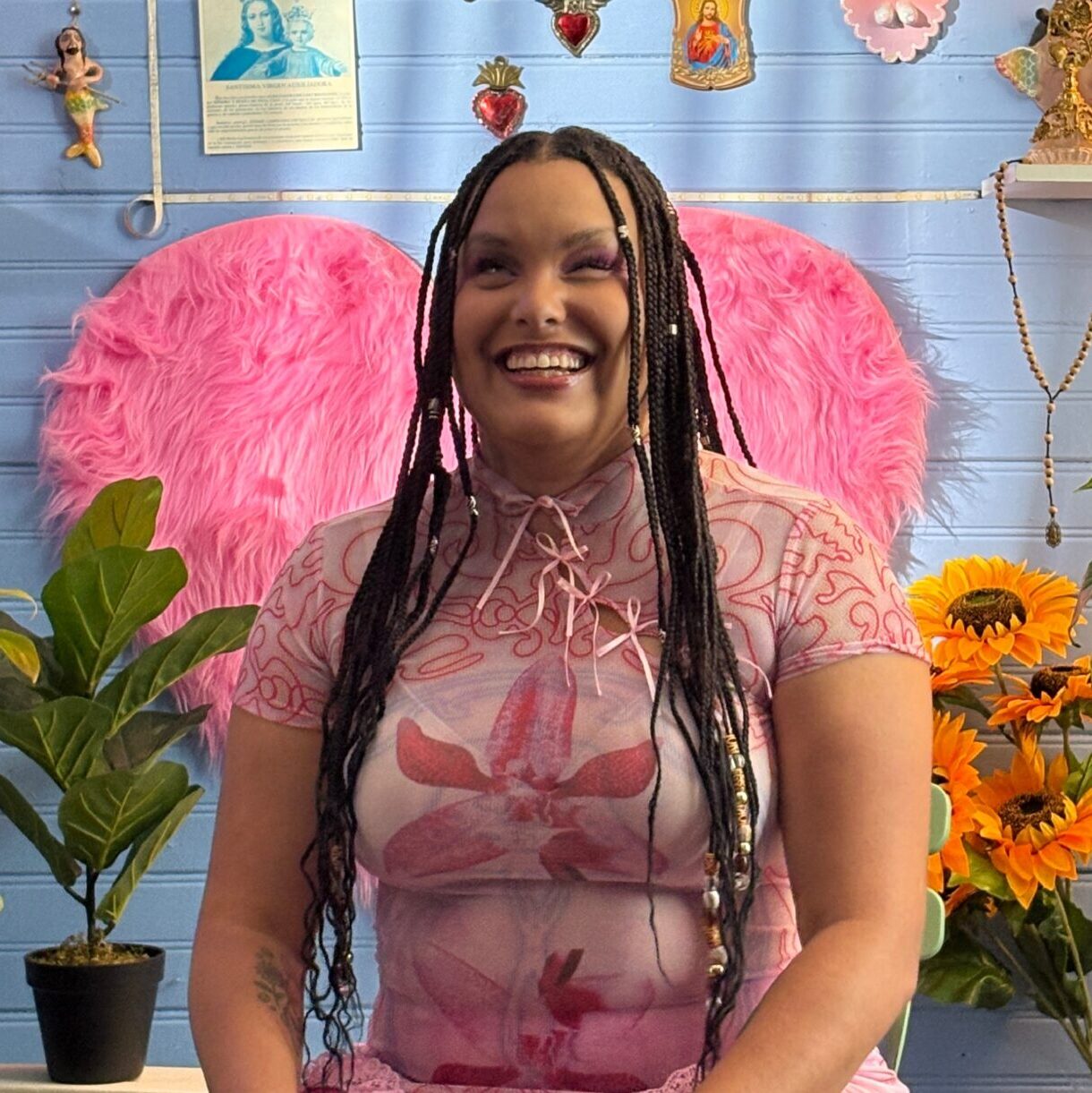 Precious sits in a room full of flowers and bright colors, wearing a soft pink tshirt. She smiles at the camera.