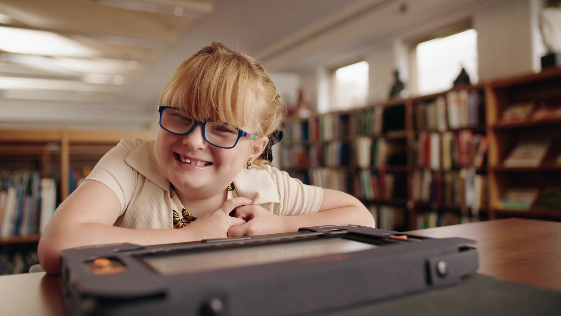 A young girl wearing glasses, butterfly earrings, and a shirt with butterflies, leans over a Monarch computer while smiling at the camera