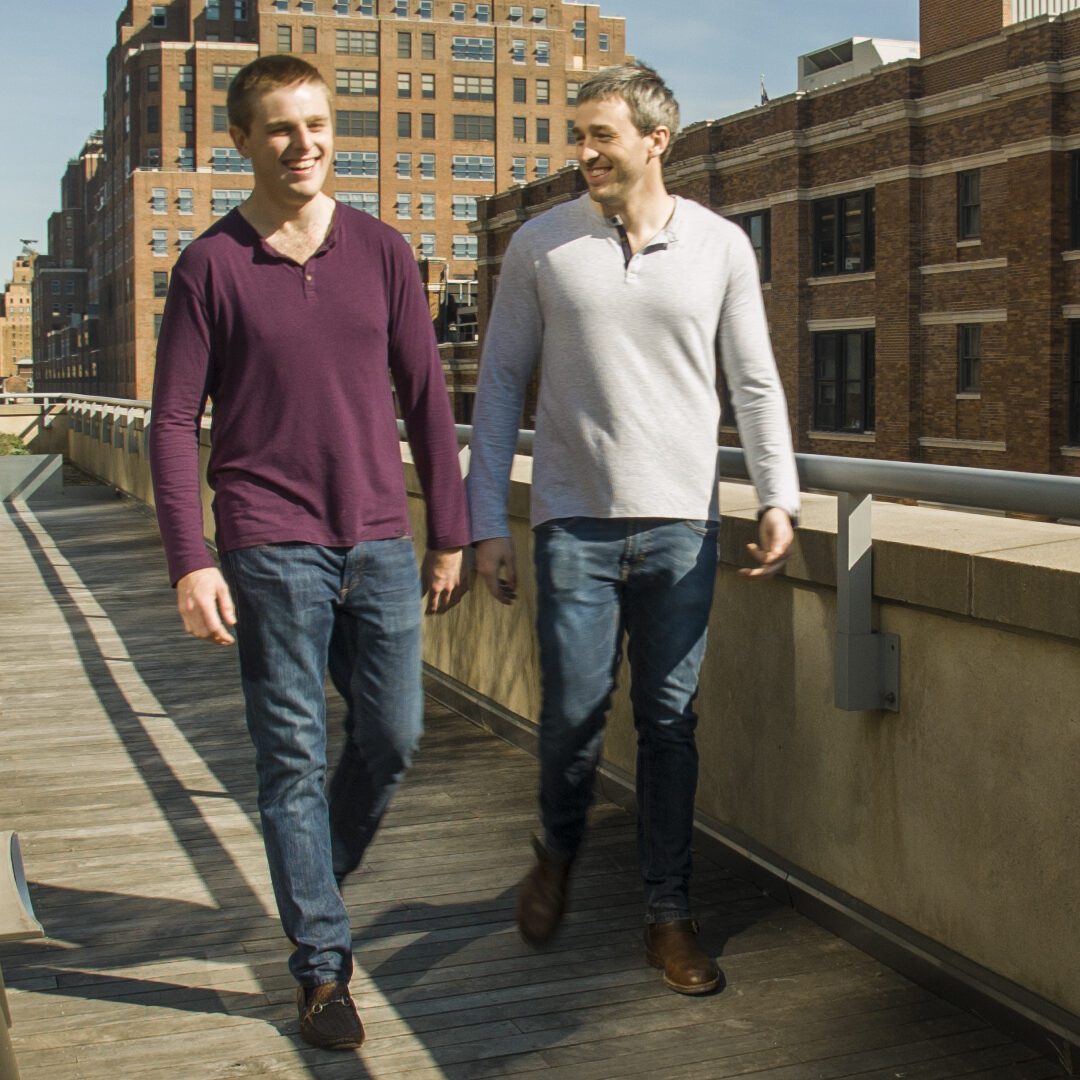 Bradford and Bryan smile and laugh as they walk side by side with tall buildings in the background.