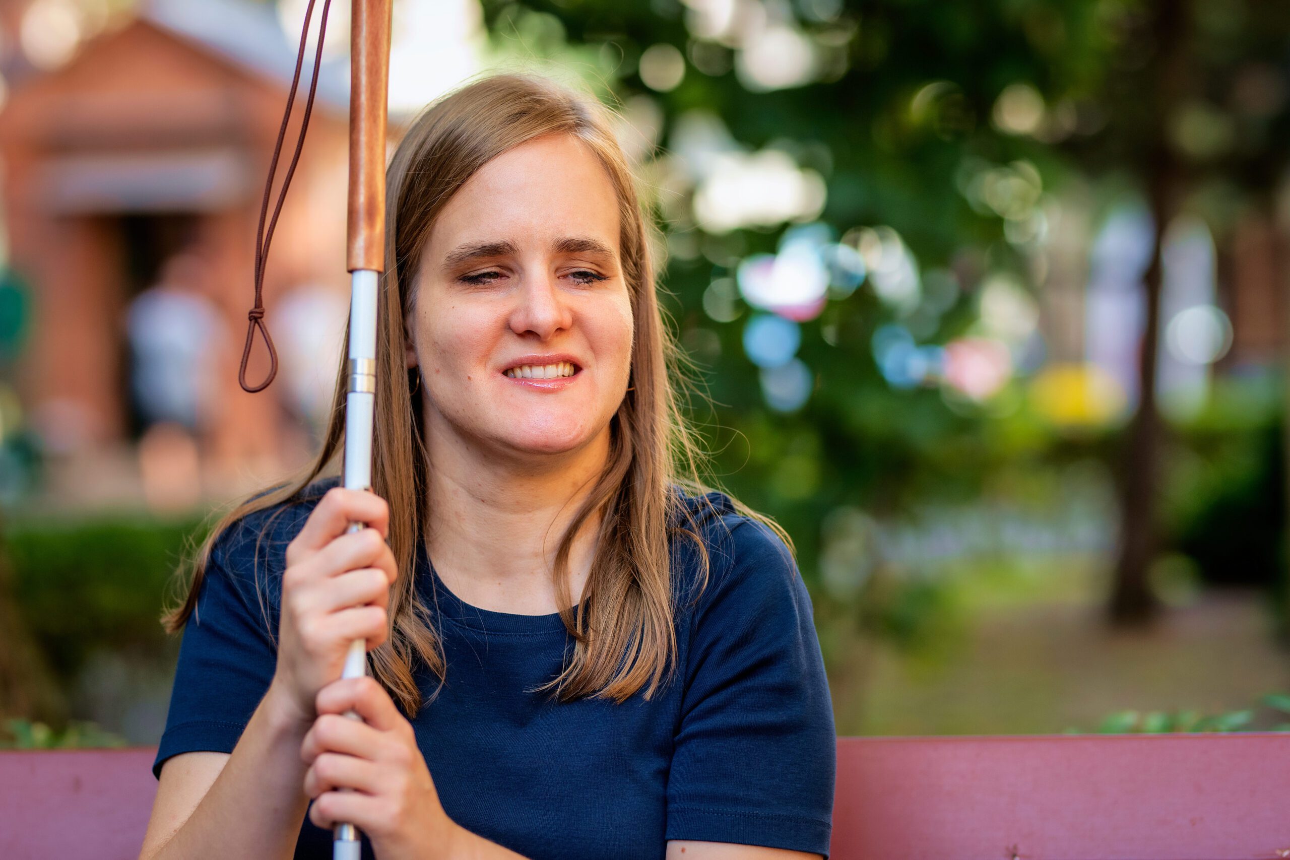 Close-up of a visually impaired woman holding a white cane and sitting on a bench in the city. Smiling female wearing casual clothes.