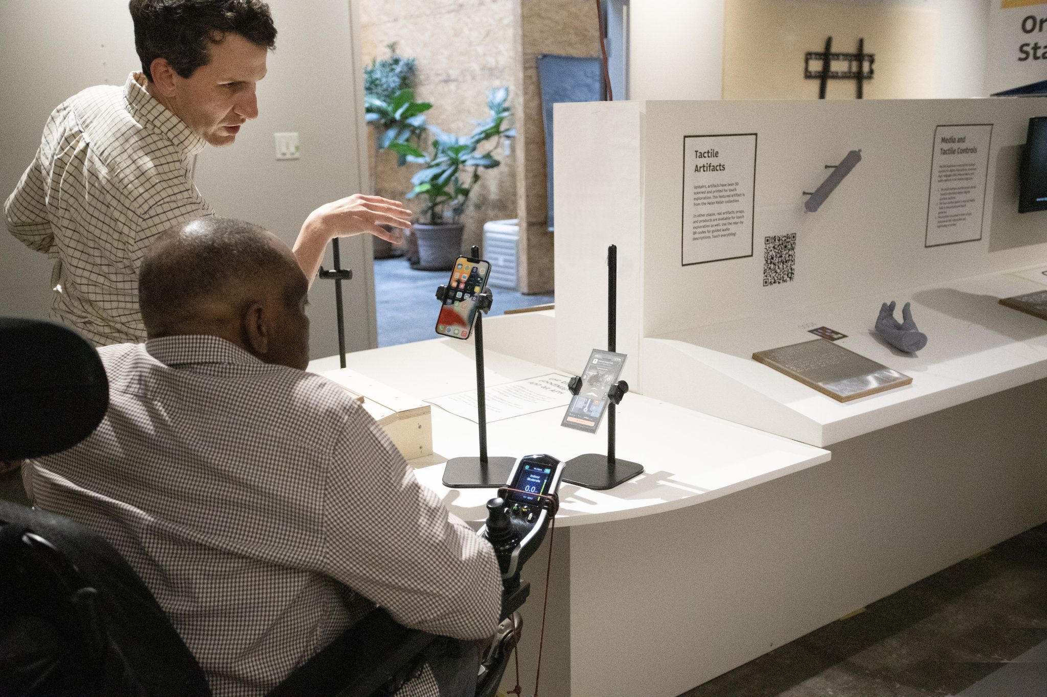 A standing man speaks to a man in a wheelchair while they both face an oval shaped table with a raised section and a short pillar in the middle. In front of the men are two black stands holding smartphones.