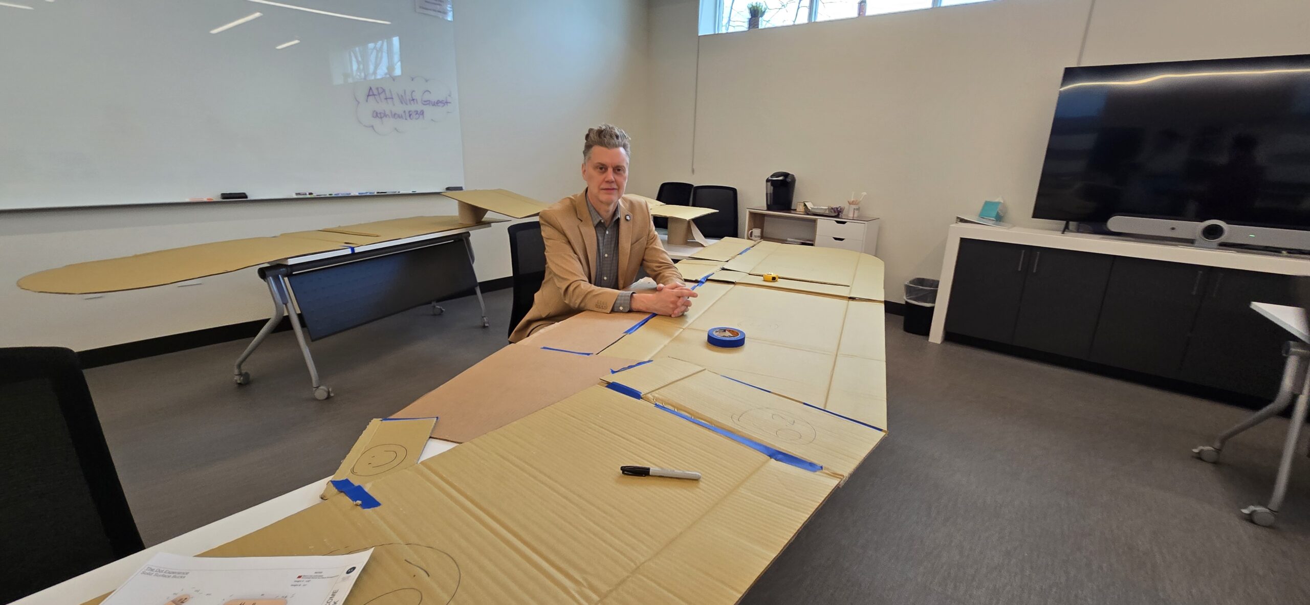 Darrick sits at a large desk in a tan suit. Across the desk are pieces of tape and cardboard.