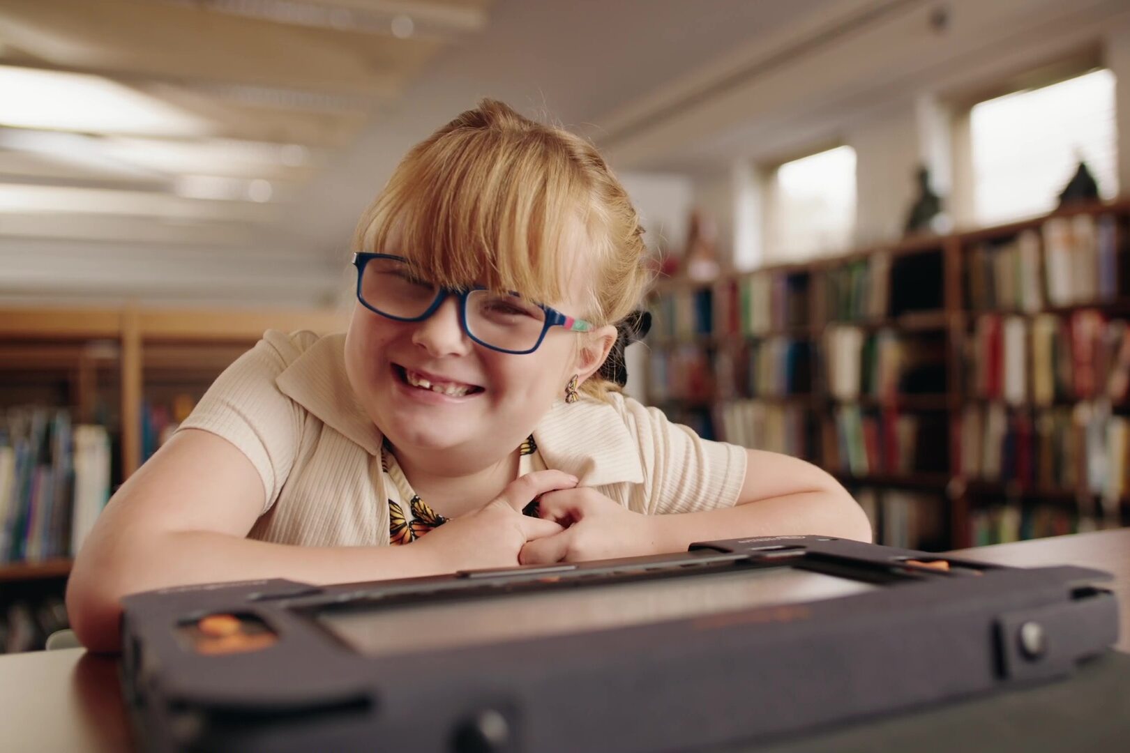 A young girl wearing glasses, butterfly earrings, and a shirt with butterflies, leans over a Monarch computer while smiling at the camera