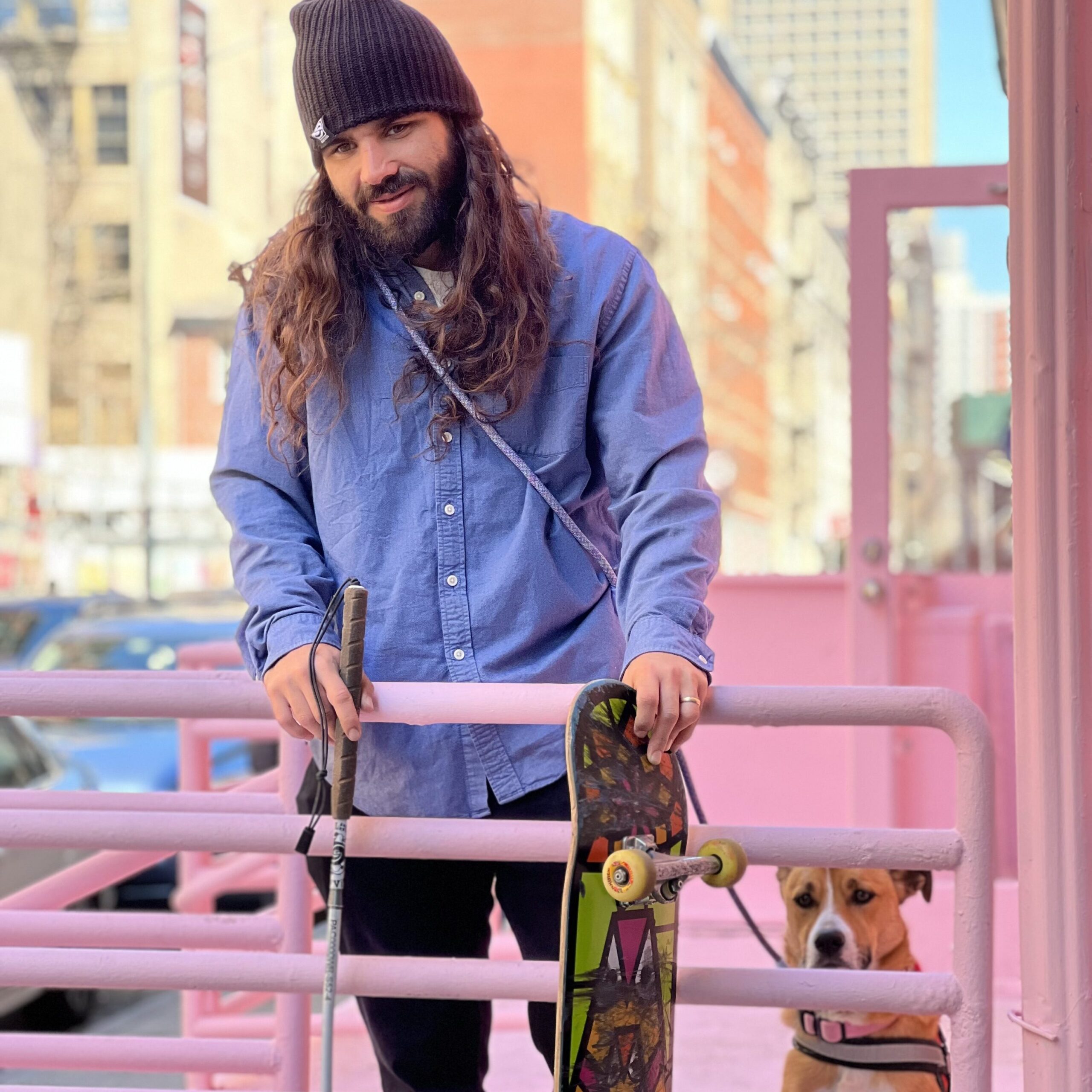 Anthony Ferraro looks back at the camera with his dog at his side. He holds a skateboard in his left hand and his cane in his right.