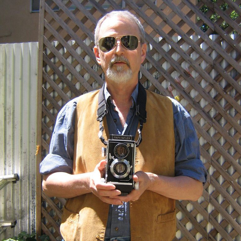 Pete stands outside wearing sunglasses and jeans with a blue dress shirt and tan vest. Facing the camera, he holds another camera in his hands that is strapped around his neck.