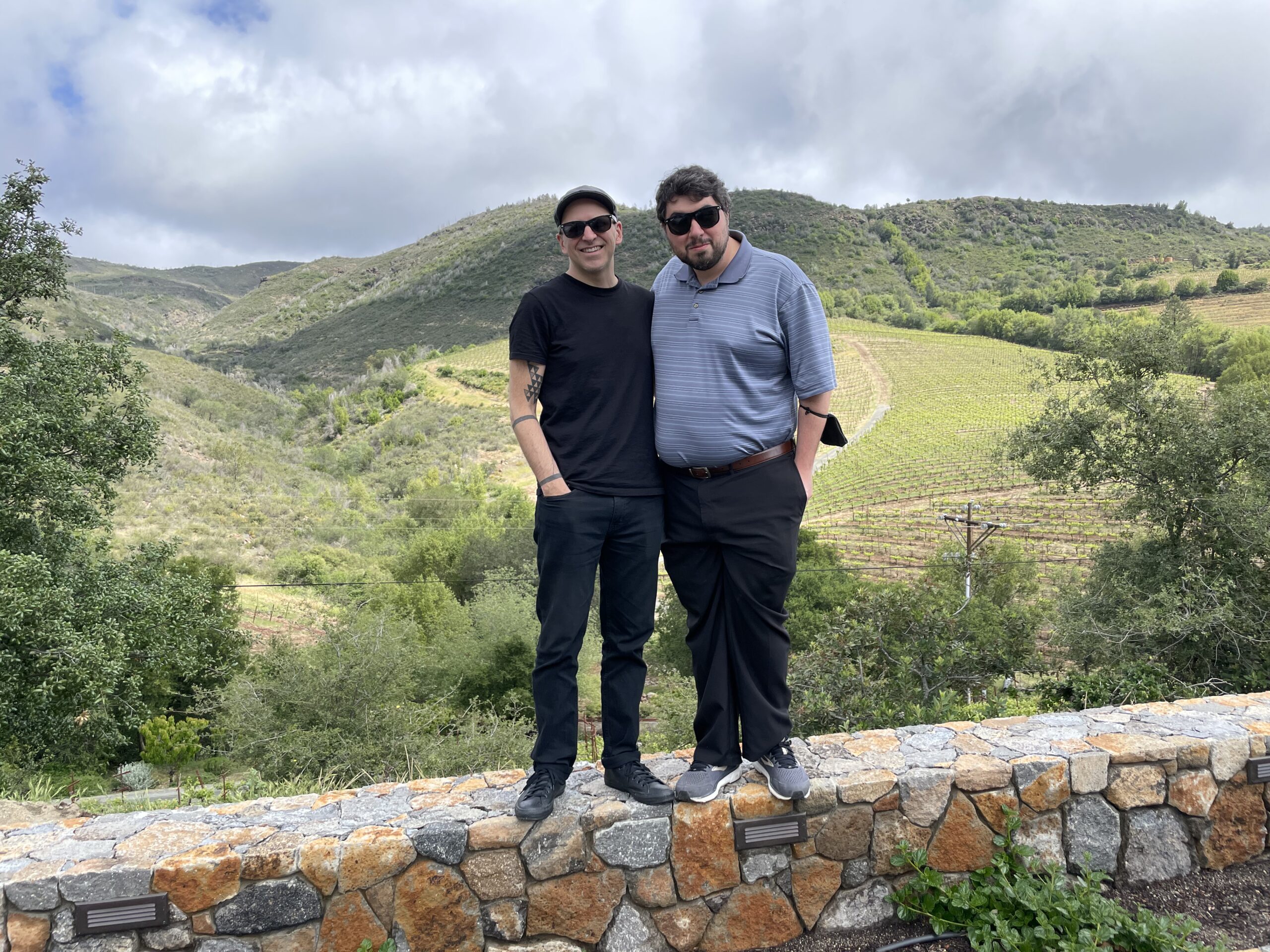 Two people wearing sunglasses pose together outdoors, standing on a low stone wall in a rural landscape. One wears a black t-shirt and pants, while the other wears a blue shirt and dark pants. They stand close together, on top of a stone wall, smiling toward the camera. Behind them, green and brown agricultural fields spread across gently rolling hills, divided into irregular patches and rows. A distant ridge runs along the horizon beneath a dramatic, heavily overcast sky filled with layers of grey clouds.