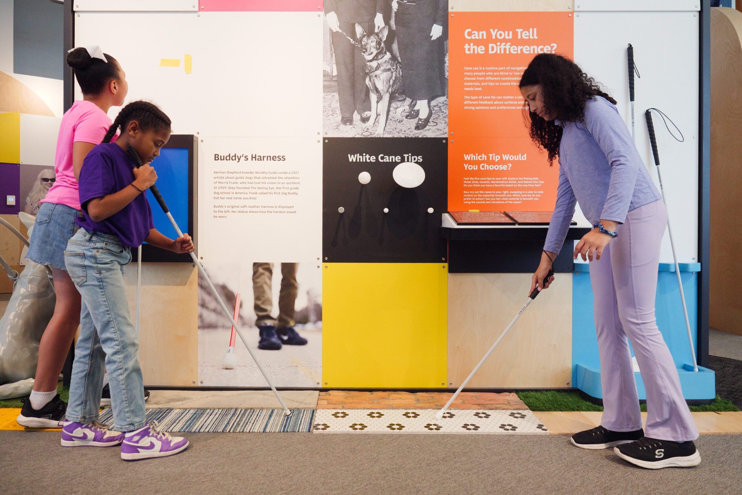 Three young girls experience what it is like to use a white cane. They are testing canes on different surfaces.