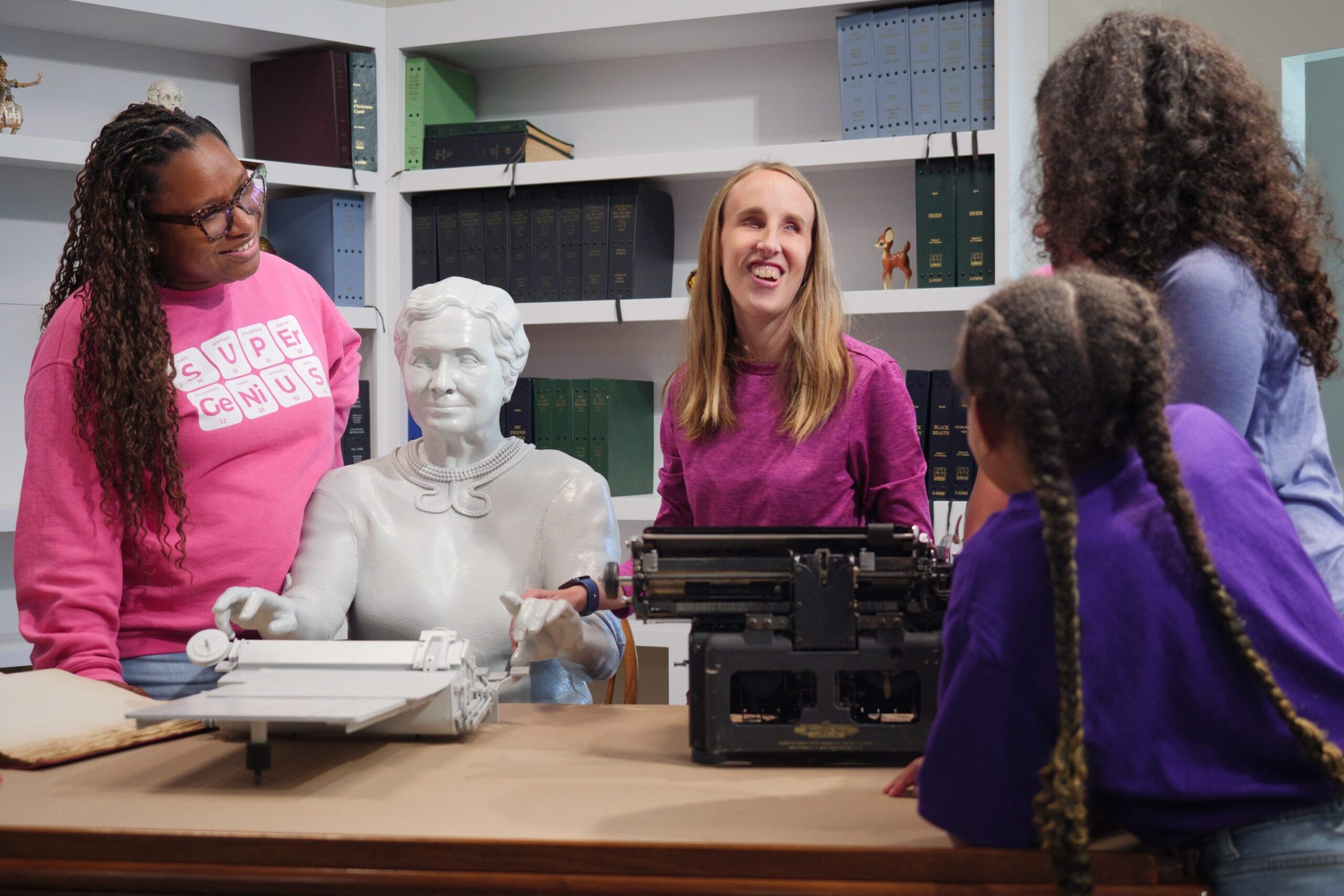Two women and several younger girls are gathered around a desk owned by Helen Keller. A life-size replica of Helen is sitting at the desk and her arms are stretched over a brailler.