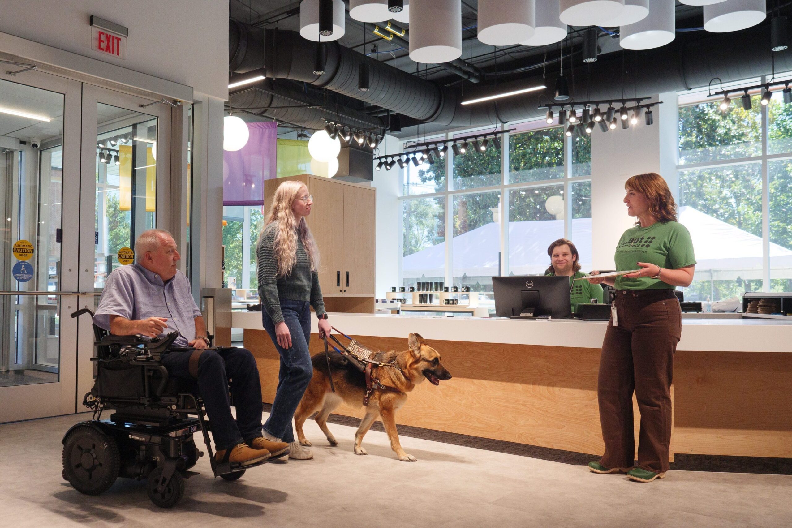 A man in a motorized wheelchair and a woman with a german shepherd guide dog are greeting in the lobby of The Dot Experience. The lobby is filled with natural light from a back wall of floor to ceiling windows. Two staff members are waiting to greet the visitors.