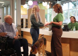 A man in a motorize wheelchair and a woman with a german shepherd guide dog in the lobby of The Dot Experience. A staff member is holding something the woman is feeling. Another staff member sits behind a welcome desk.