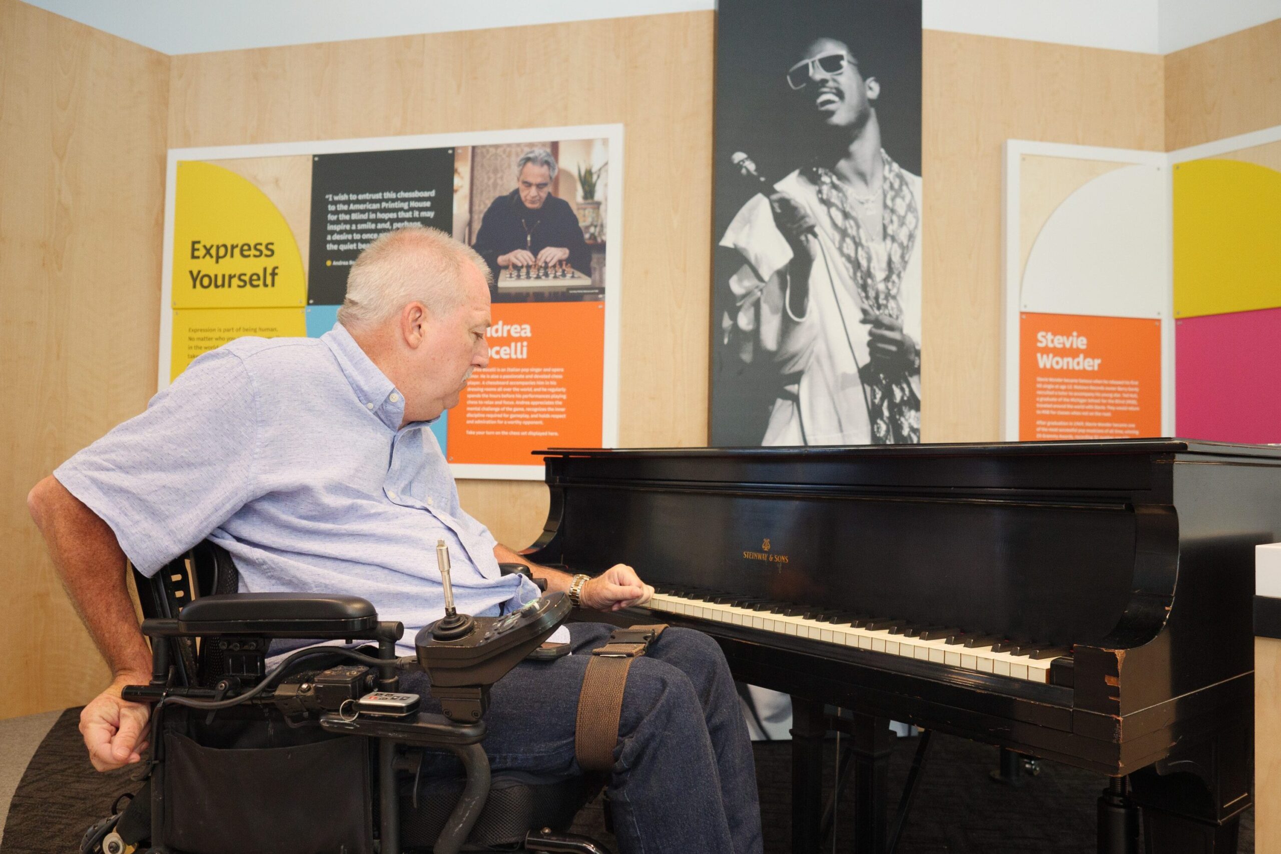 A man in a motorized wheelchair is looking at a baby grand piano. His left hand is resting on the keys. Behind the piano is a large photo of Stevie singing and holding a microphone.