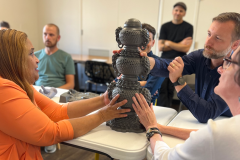 Three individuals sit around a white folding table. They are touching a 3D-printed, plastic, three-tiered incense burner that is sitting on the table in front of them. The incense burner is grayish black in color. The base of the incense burner is shaped like a loaf of bread that resembles a pile of grayish black coins. On top of the base sits one chamber of the incense burner. This chamber is shaped like a rounded jar, complete with two ornate handles that resemble seahorses with manes on either side of the jar. Atop this section of the burner sits three rows of the coin-style texture featured at the bottom of the structure. Atop those coins sits a smaller yet identical rounded jar with the same handles. This pattern is repeated one more time above in a smaller iteration to create the third level of the burner. The structure is topped with a small round dome reminiscent of the speaking piece on a microphone. The entire burner is covered in numerous small indentations roughly 1 cm in diameter each. The woman on the left has strawberry blonde hair and a medium complexion. She is wearing an orange shirt and is touching the largest jar with two hands. In the bottom right-hand corner of the image, a white woman with brown hair is wearing a white blouse, glasses, turquoise earrings, a watch with a black leather band, and two cloth bracelets. She has one hand on the largest jar and one hand at the base of the incense burner. A white man sits in the top right corner of the image. His right hand is grasping the burner between the middle and top levels. He is wearing a dark blue button-up shirt, a gold wedding band, and a digital watch with a cloth band. He has a salt and pepper beard and light brown hair that has been parted and combed with gel. Three people in the background sit out of frame and longways down the white folding table. A man in a black shirt and newsboy hat stands in the background with his arms folded, observing the object interaction.