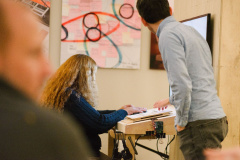 A woman sits at a kiosk with hands touching a refreshable braille device that sits in front of a monitor. To the right of the image a designer is reaching forward and offering guidance to the person operating the station.