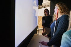 A person with a cane is standing close to a screen and smiling. Standing in the distance is a designer observing and evaluating, and also smiling.